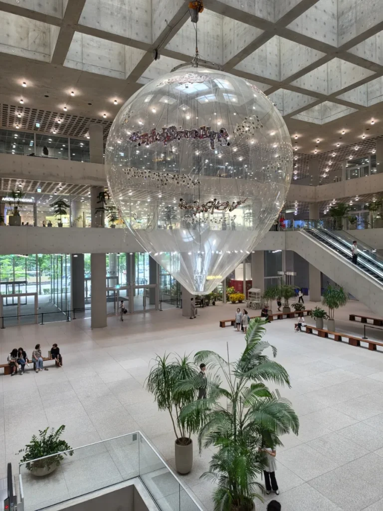 Large transparent installation suspended above benches and plants beneath the concrete grid ceiling of the Amorepacific atrium.