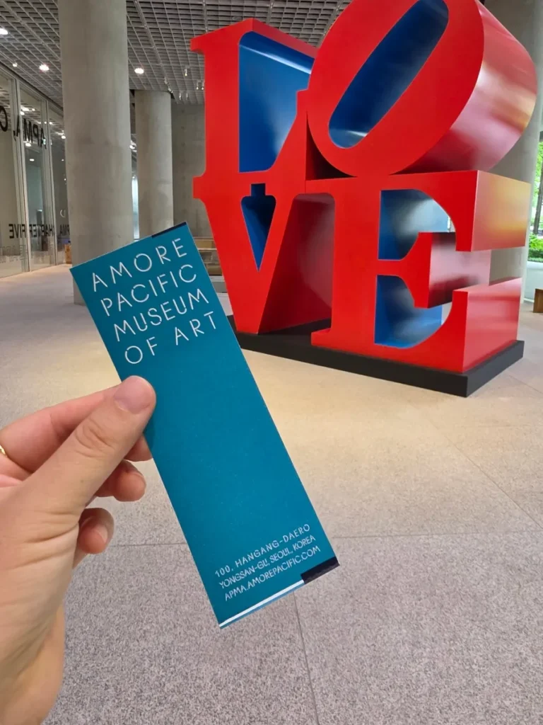 Hand holding an Amorepacific Museum of Art ticket in front of a red and blue LOVE sculpture in Seoul.
