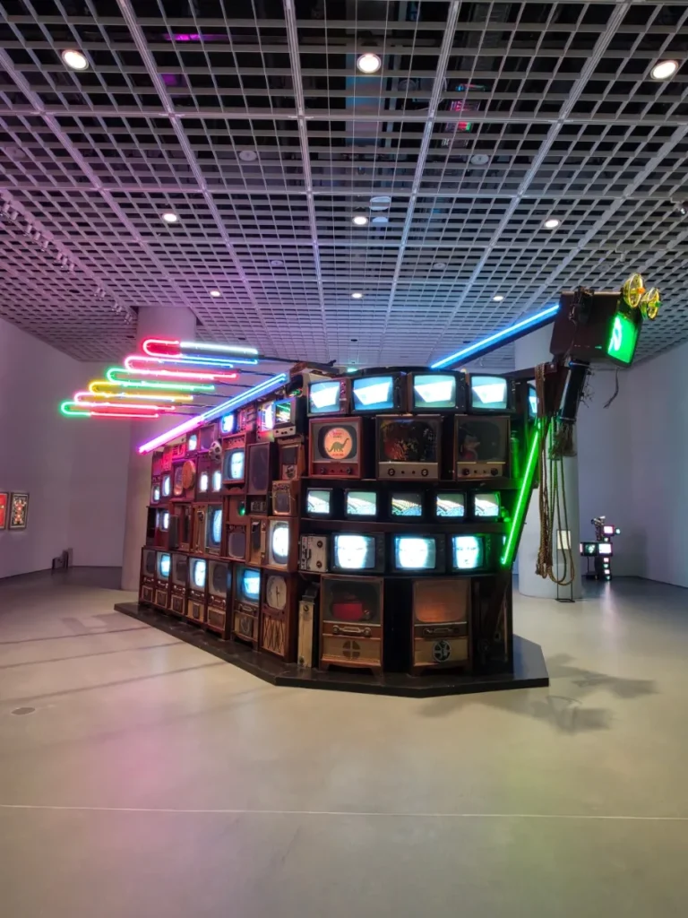 Large Paik Nam June installation combining stacked television sets with pink, blue, green and yellow neon lights.