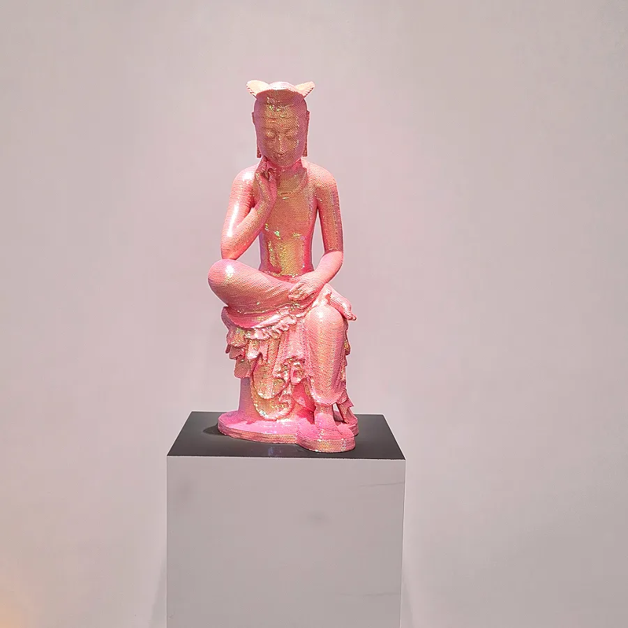 Pink sequin-covered Buddhist figure seated with one leg raised and a hand near its cheek on a museum pedestal.