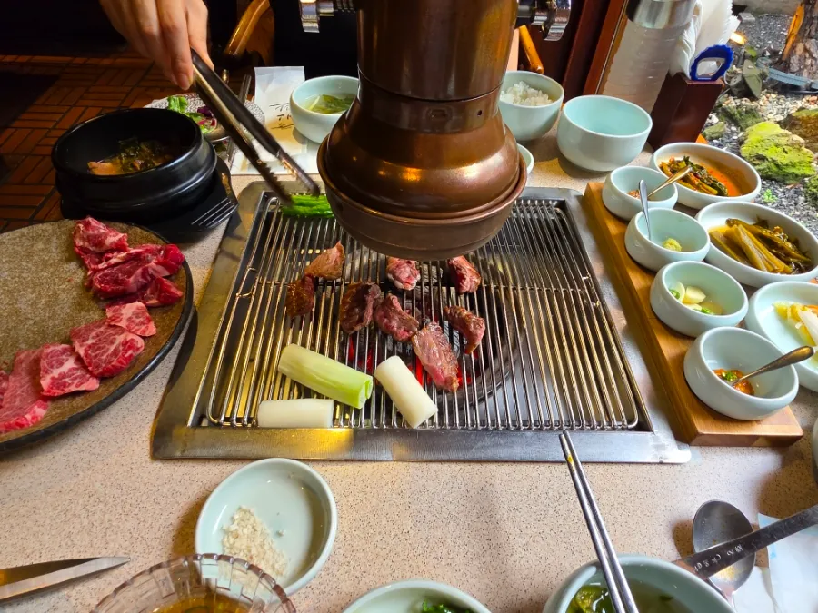 Hanwoo beef and vegetables cooking on a tabletop grill surrounded by Korean side dishes.