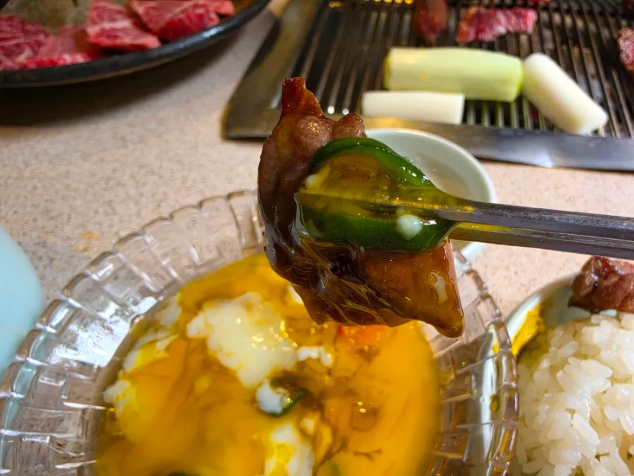 Grilled beef held with chopsticks above a dipping sauce containing soft egg and sliced green chili.